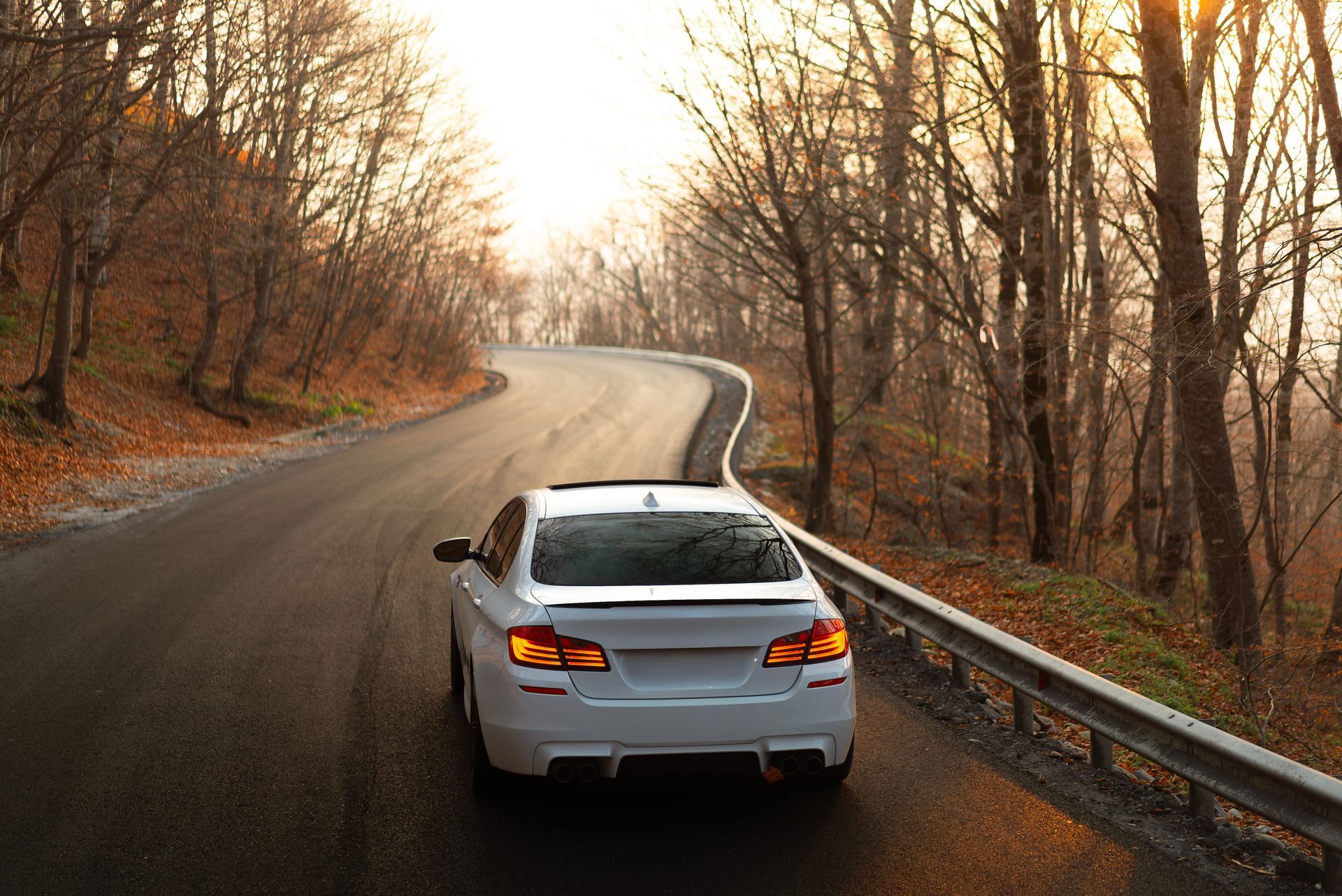 White car driving on a winding forest road in autumn with bare trees and warm sunset light. White car driving on a winding forest road in autumn with bare trees and warm sunset light.