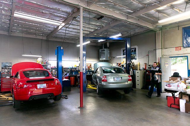 Inside auto repair shop: red car up on lift, gray car on lift, mechanics working.