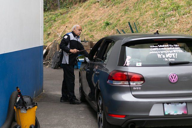 Man in uniform leans into a gray car, possibly at a gate. Blue and white building, yellow vacuum, hill in background.