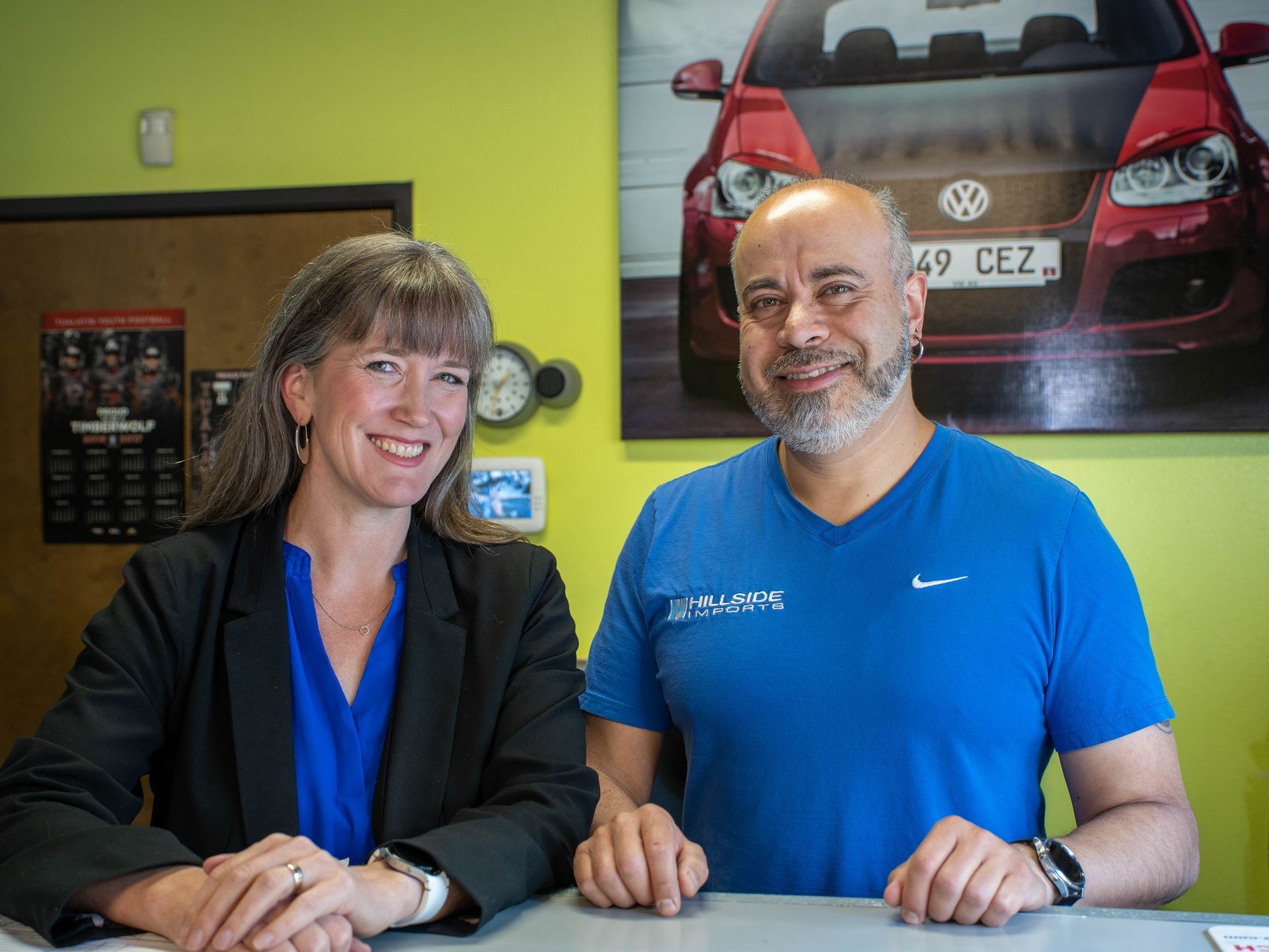 Two people standing at a counter; woman in a blazer and man in a blue shirt smiling near a car advertisement.