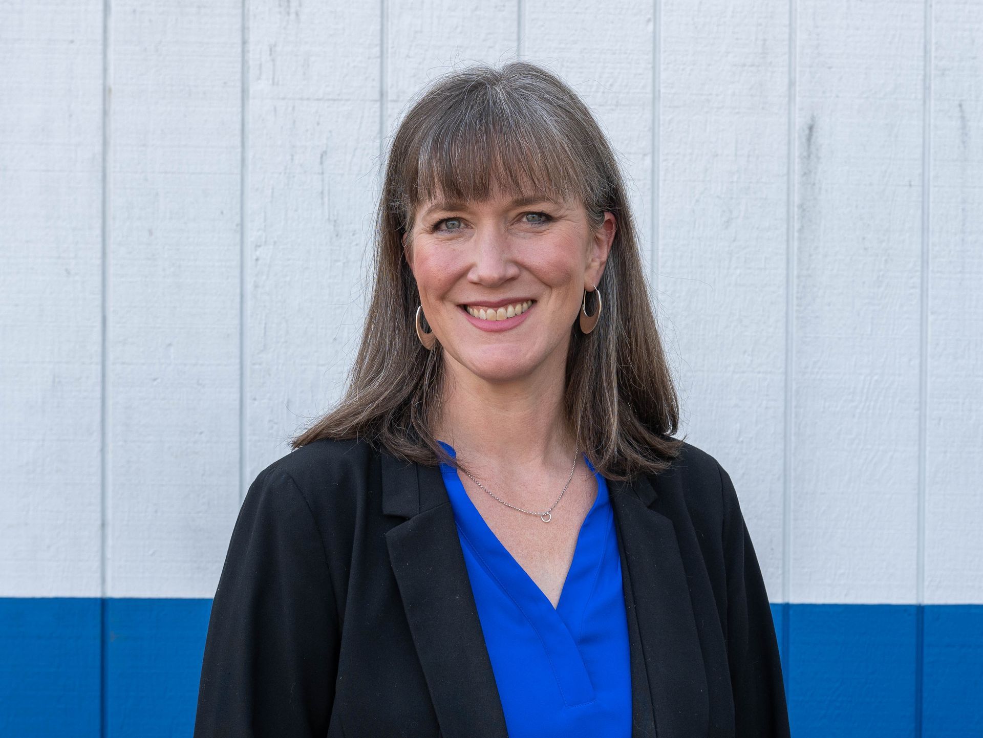 Woman in blue shirt and black blazer smiling in front of a white and blue wall.