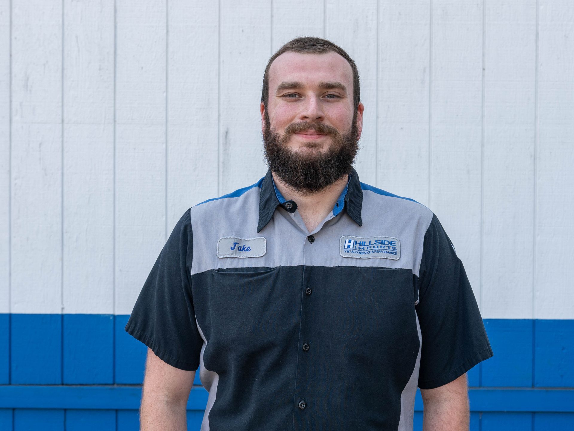 Man in blue and gray work shirt smiles outdoors in front of a white and blue wall.