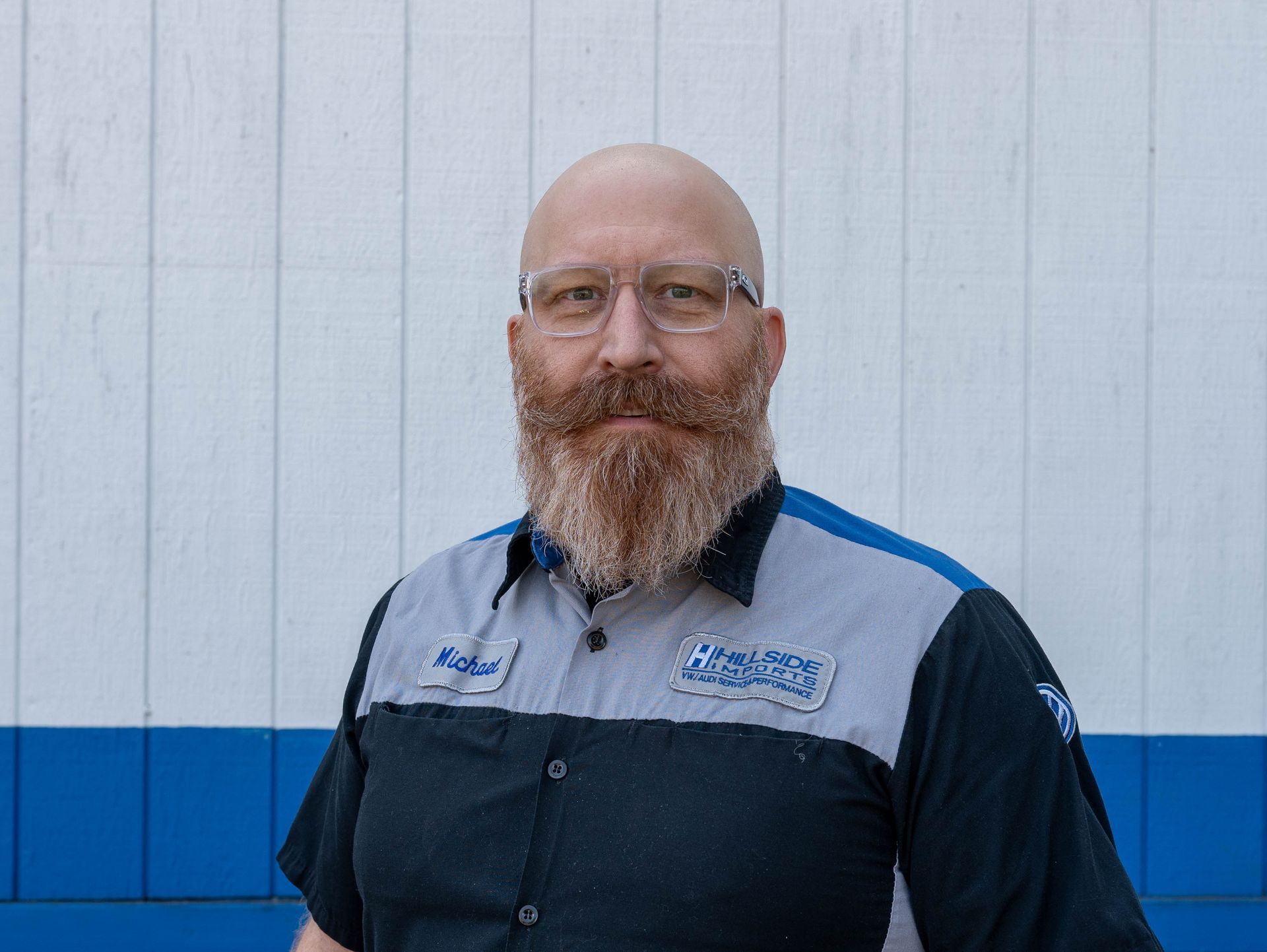 Man with glasses and beard, wearing a mechanic uniform, standing in front of a blue and white building.
