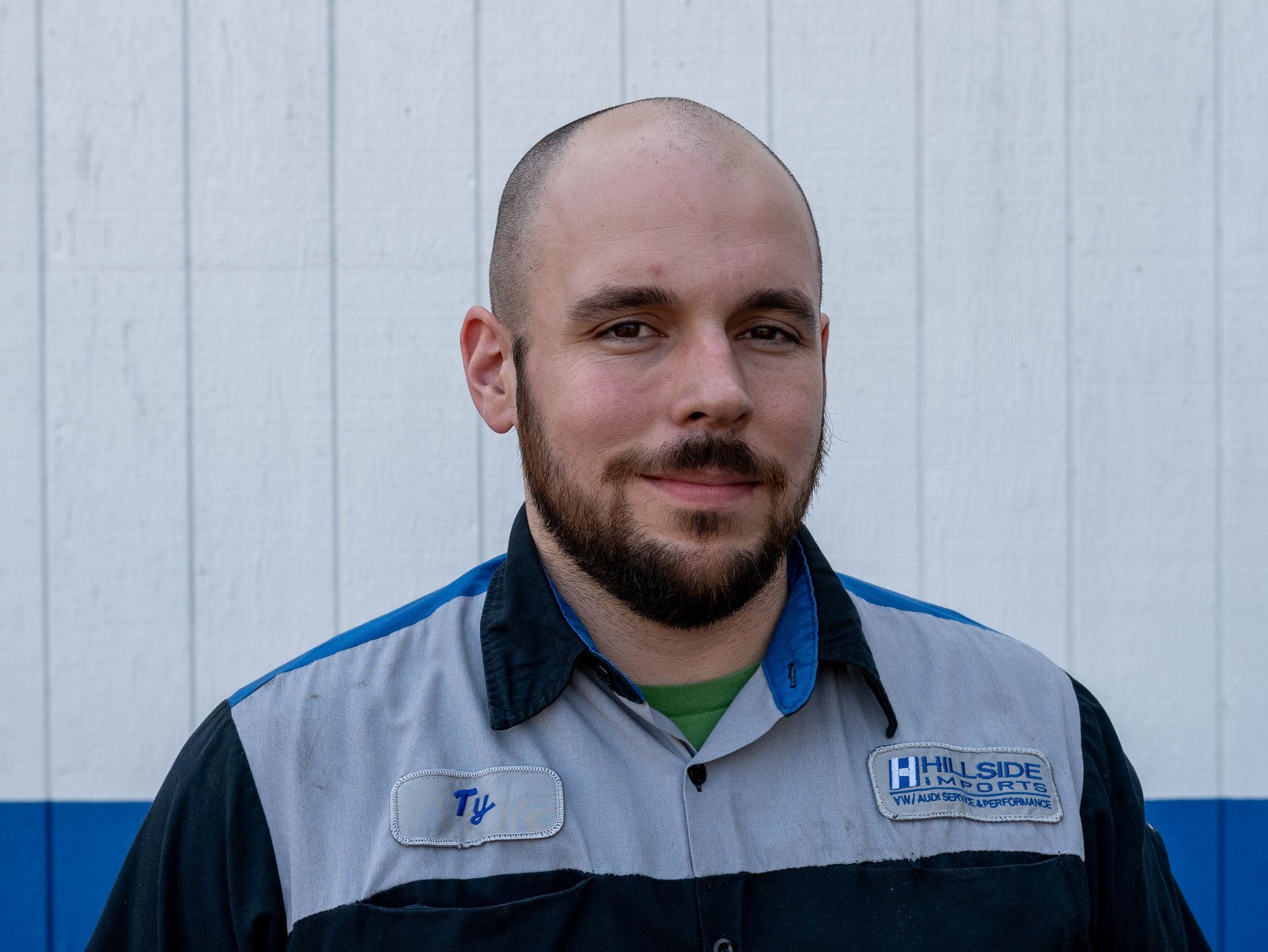 Man with a beard in a blue and gray uniform smiles in front of a white building with blue trim.