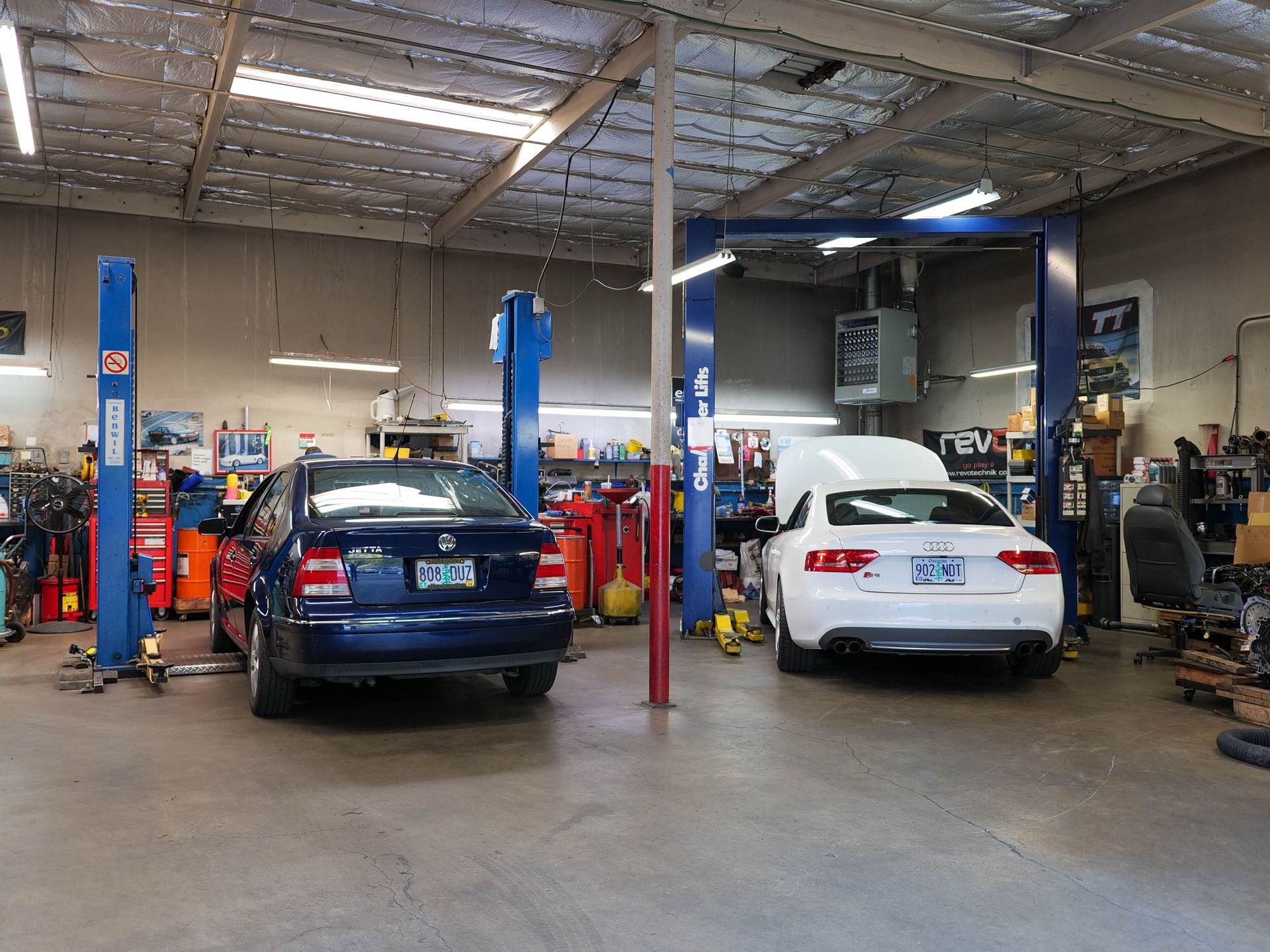 Cars in a mechanic's shop: blue sedan on a lift, white car with open hood, tools, and equipment.