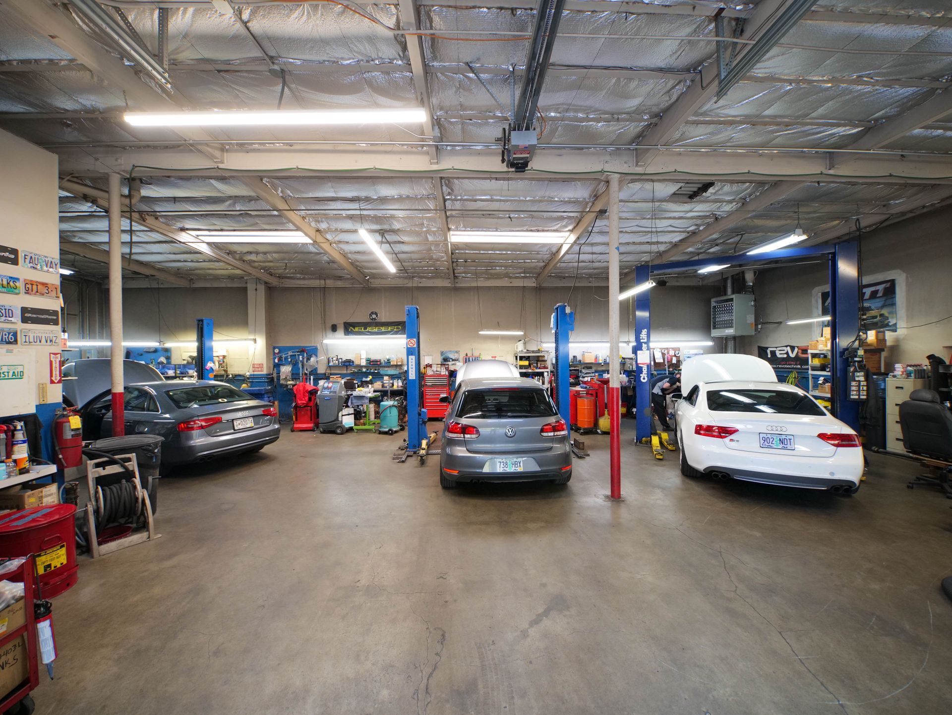 Cars on lifts inside an auto repair shop. Fluorescent lights illuminate the gray concrete floor.
