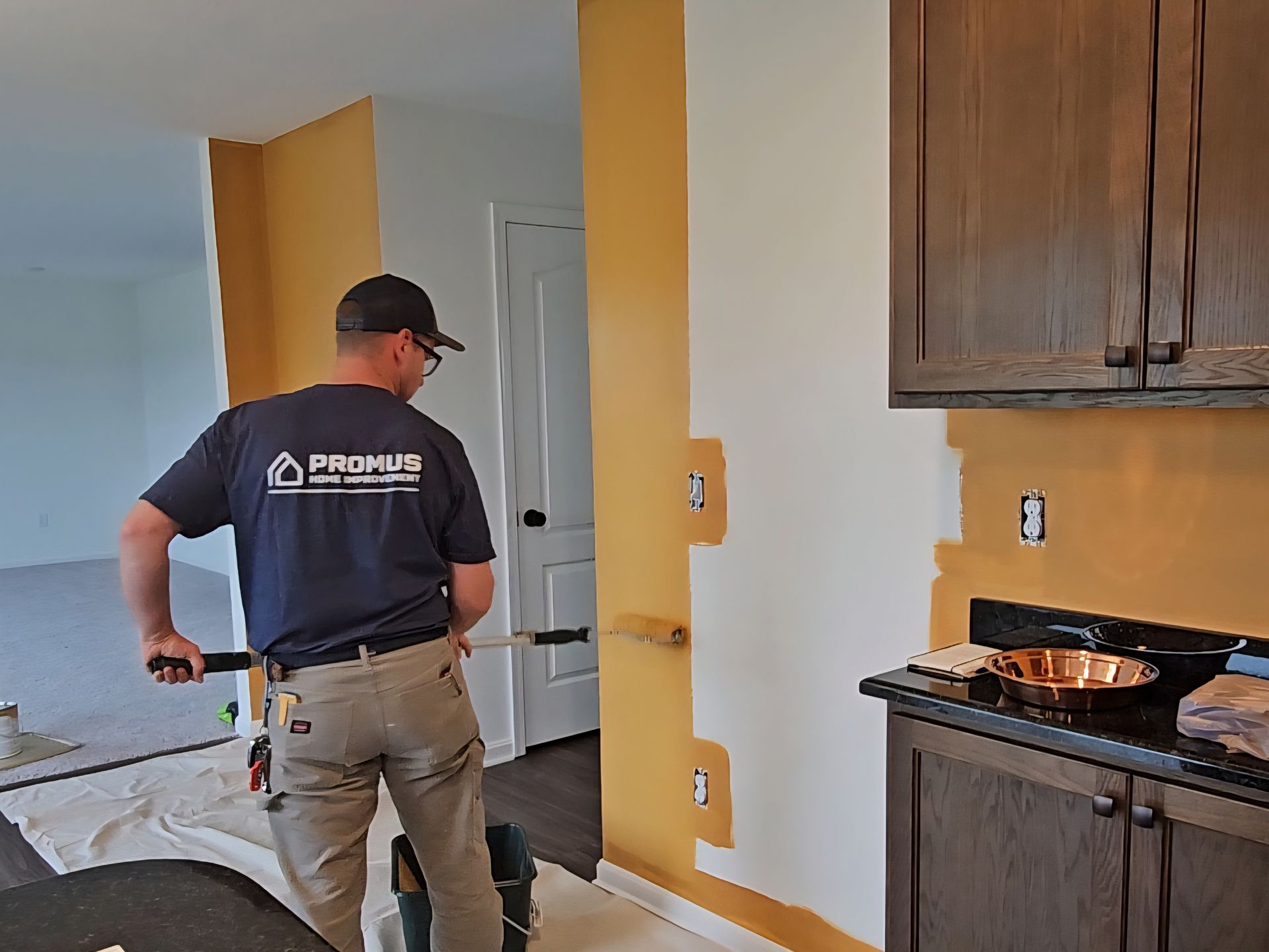 Painter in a navy shirt and tan pants painting a wall yellow in a kitchen. Powel interior painting project. 