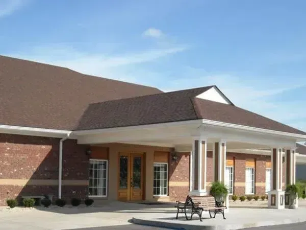Brick building exterior with white columns, brown roof, double doors, and benches in front.