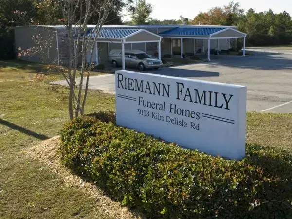 Riemann Family Funeral Homes sign in front of a white building with carport and lawn