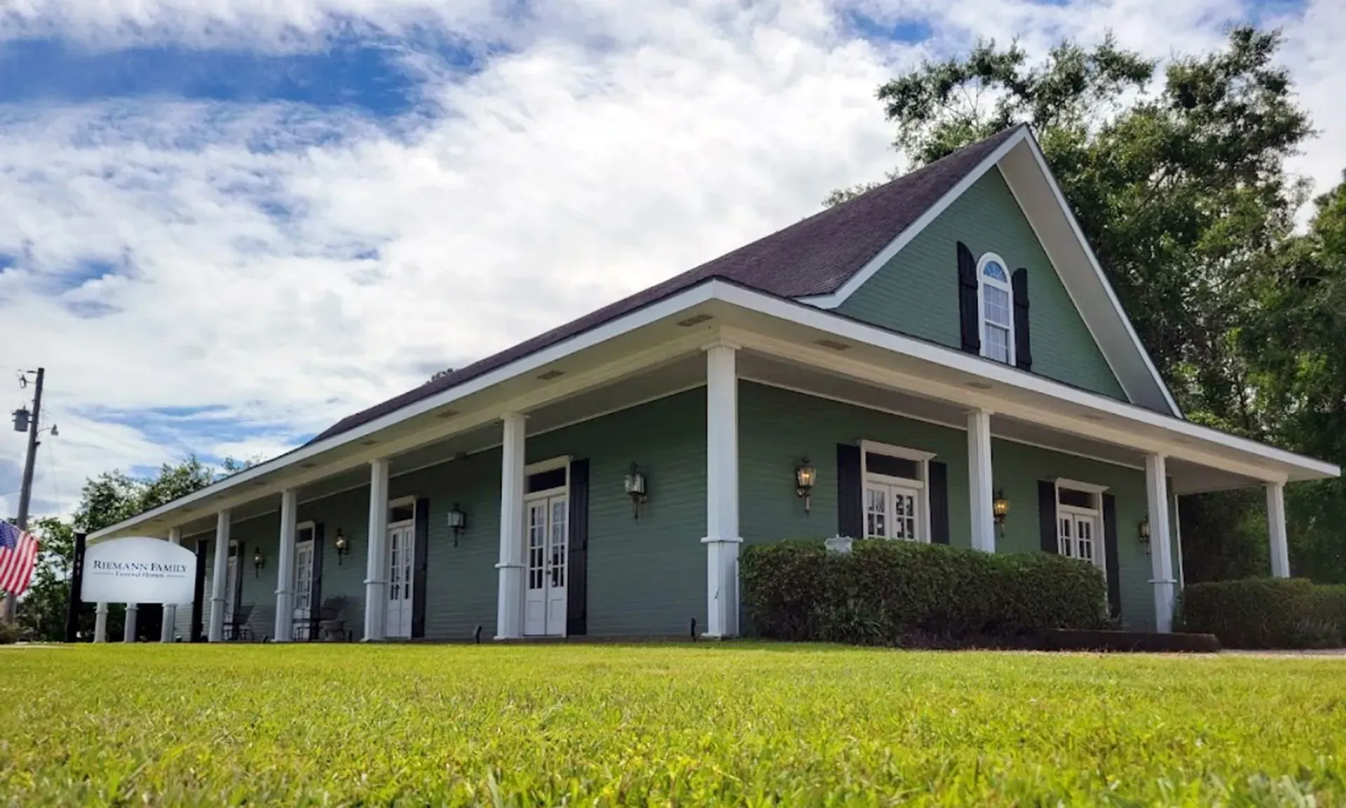Green building with white trim and wraparound porch, seen from a grassy lawn under a blue sky
