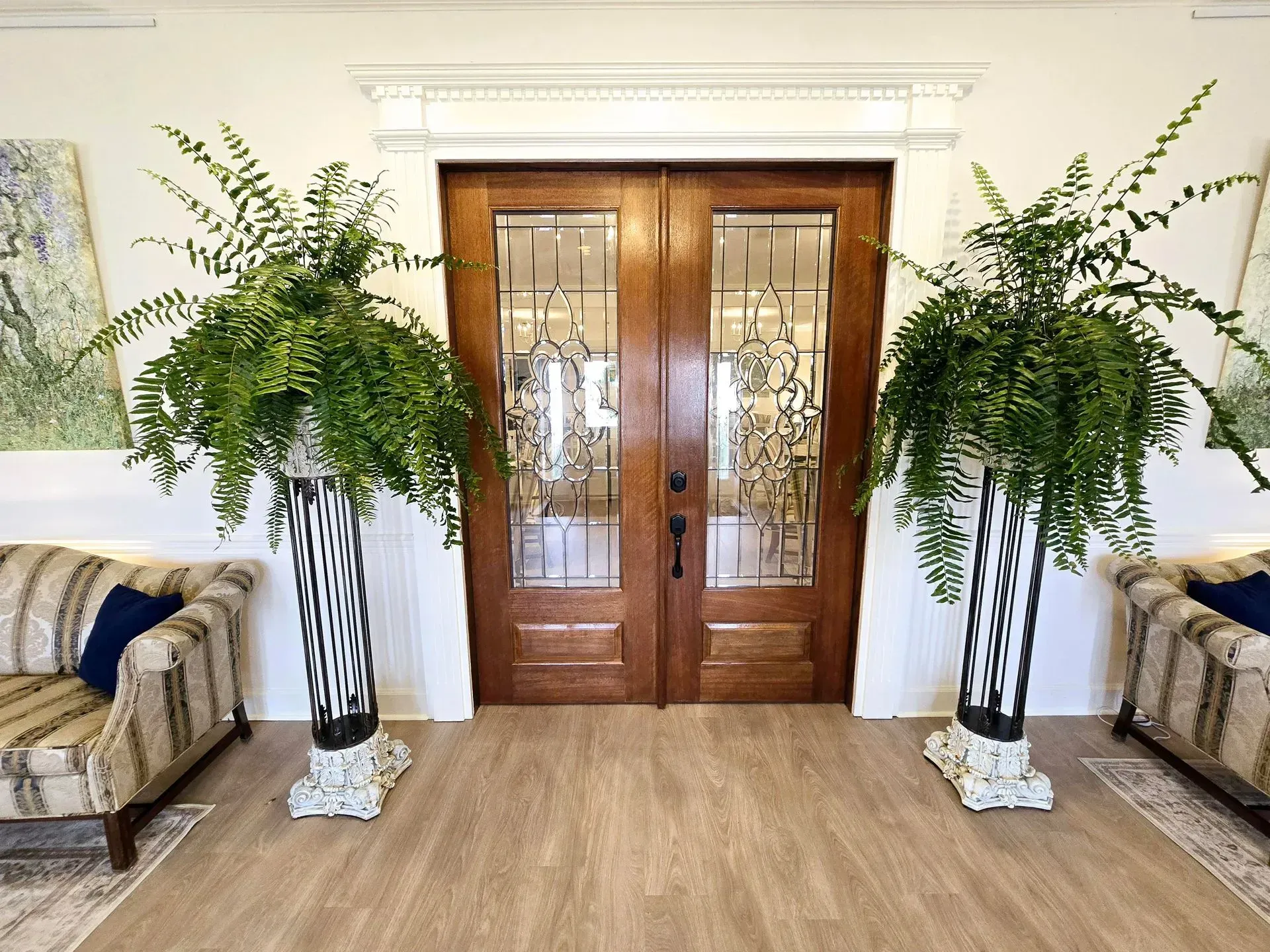 Elegant entryway with wooden double doors, mirrored sofas, and tall potted ferns on either side.