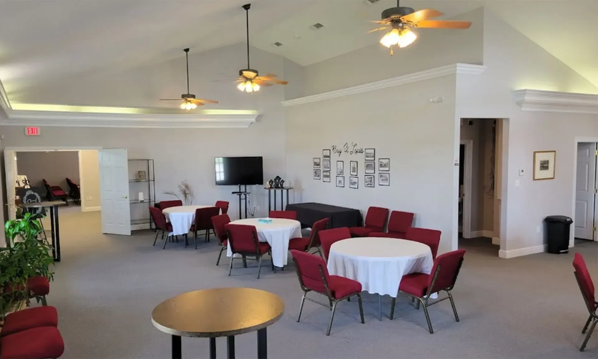 Spacious banquet room with round tables, red chairs, ceiling fans, and a TV near the back wall.
