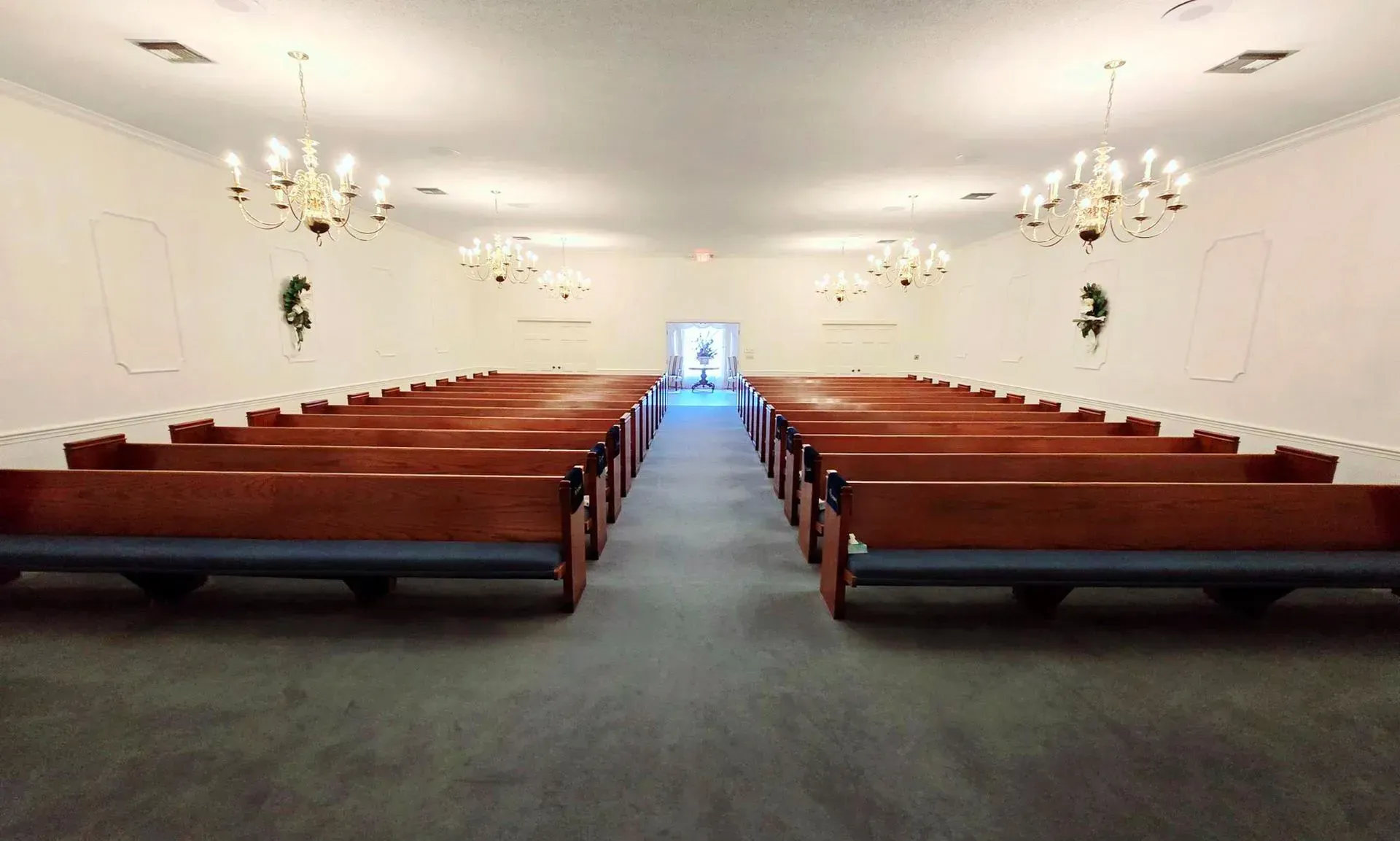Interior of a white chapel with wooden pews, chandeliers, and a central aisle leading to a bright doorway