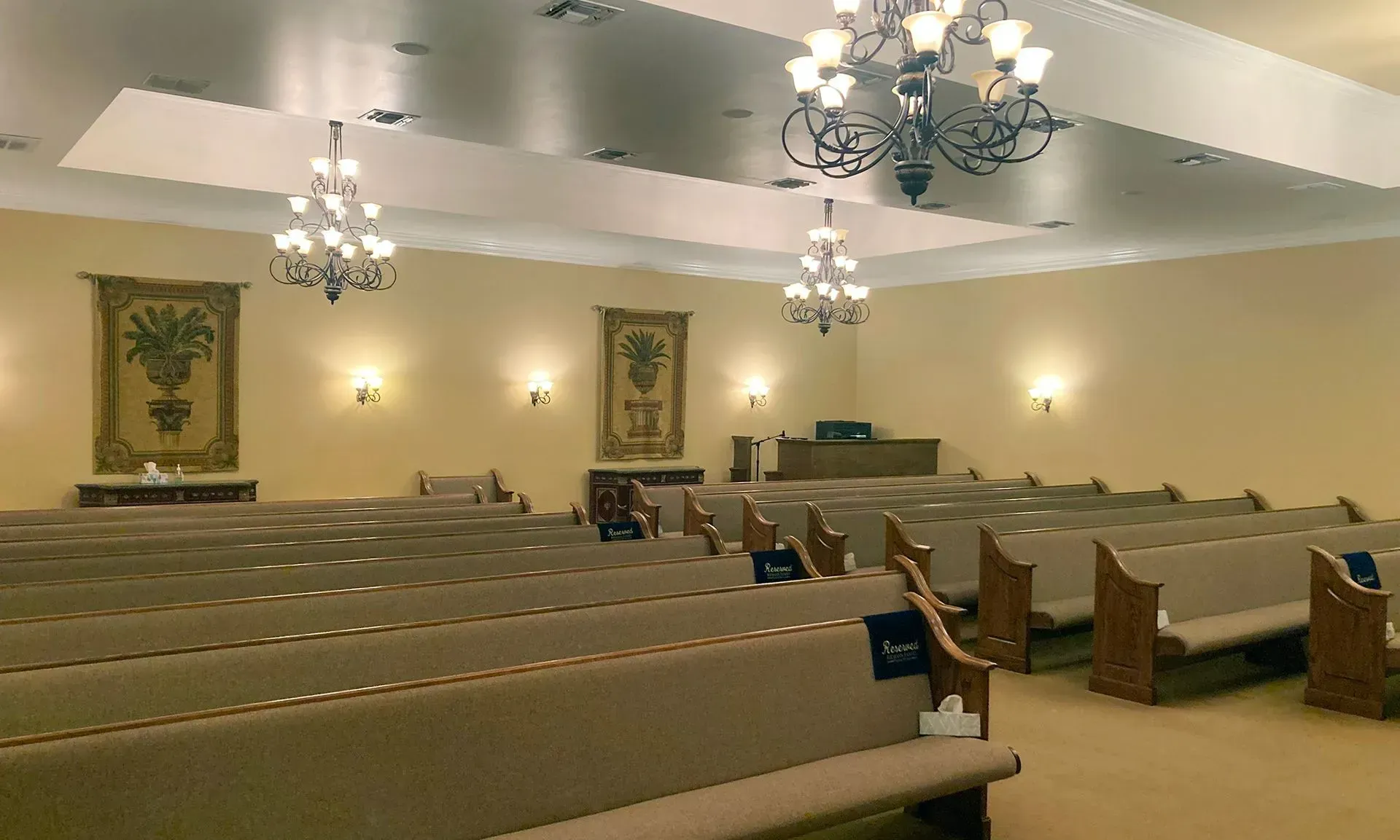 Empty church sanctuary with rows of beige pews, chandeliers, and framed wall art under warm lighting