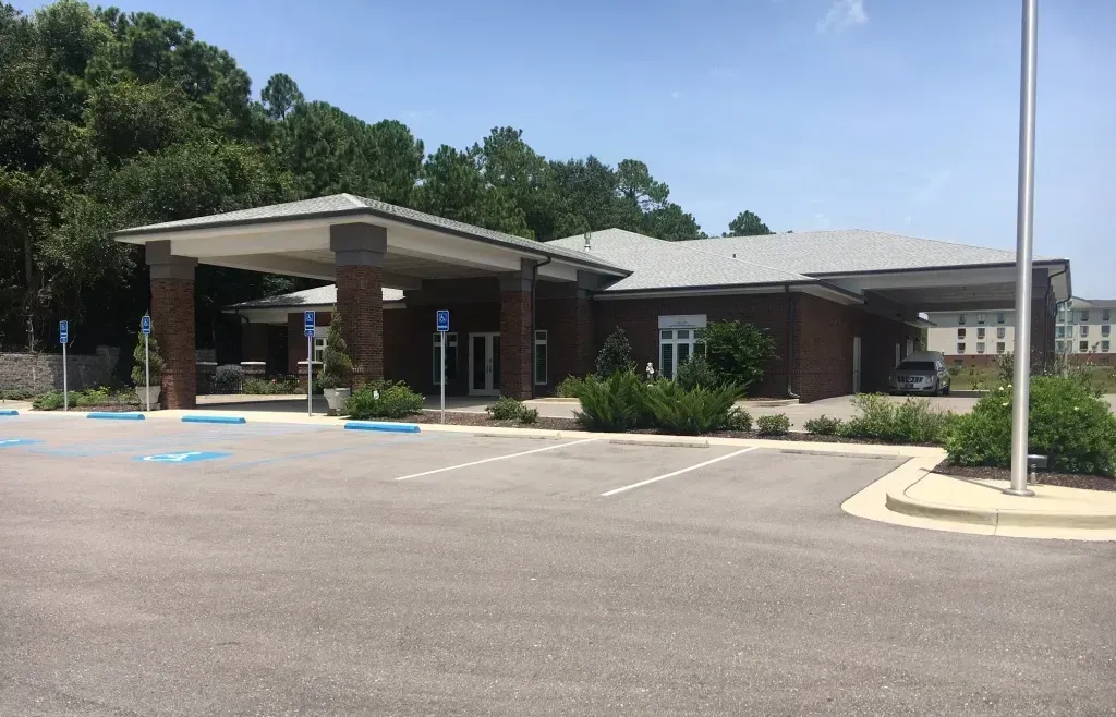 Single-story public building with covered entrance, parking lot, and trees on a sunny day