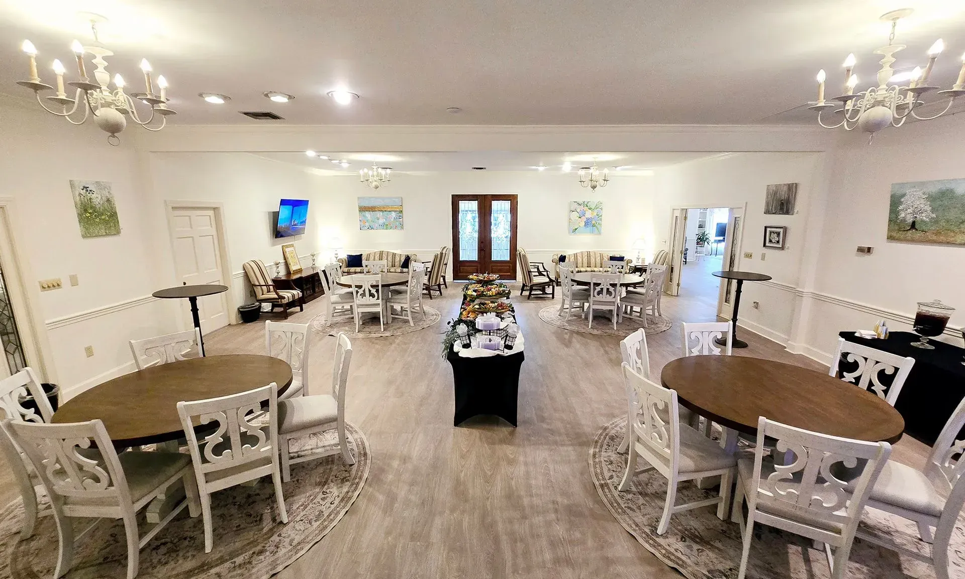 Bright dining hall with white tables and chairs, chandeliers, and a central serving table on wood floors