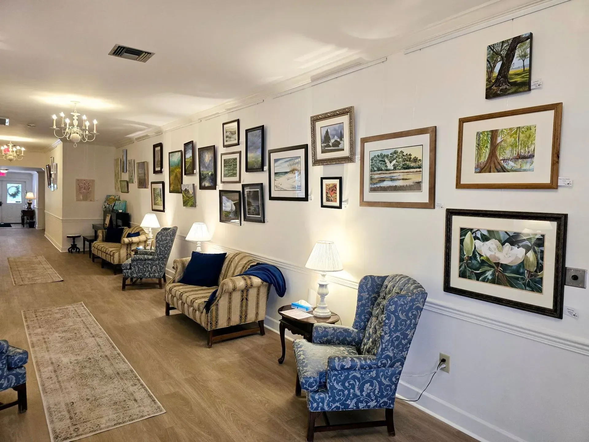 Long hallway with framed artwork, patterned chairs, side tables, and lamps in a softly lit waiting area.