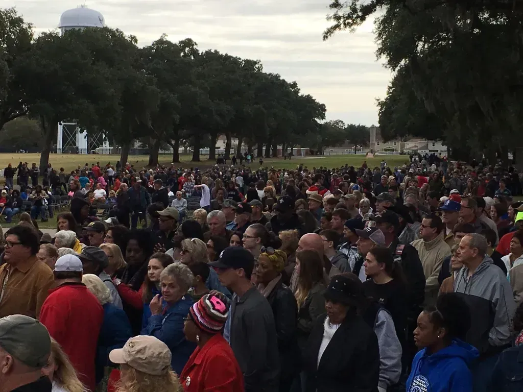 Large crowd gathered outdoors in a park, with trees and a cloudy sky in the background.