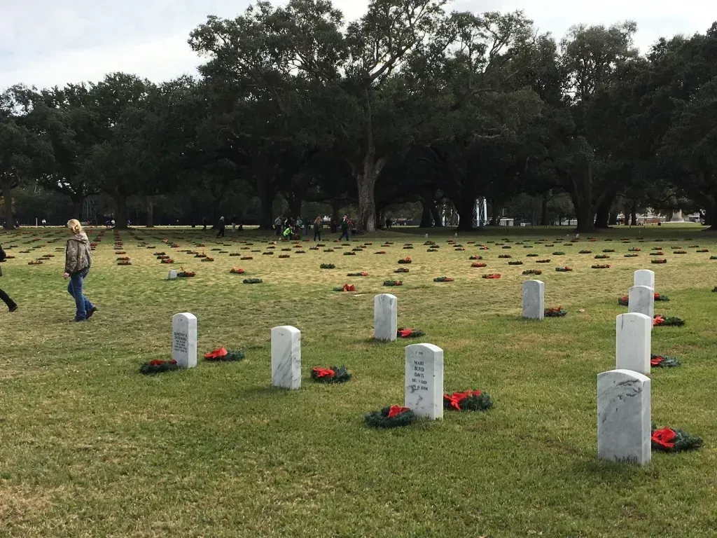 Grassy memorial field with white grave markers and red wreaths under trees; one person stands at left.