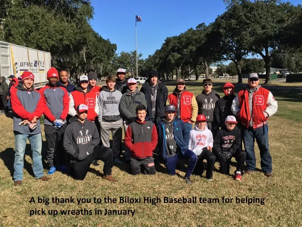 Group of baseball team members in red jackets posing outdoors with a thank-you sign and American flag behind them