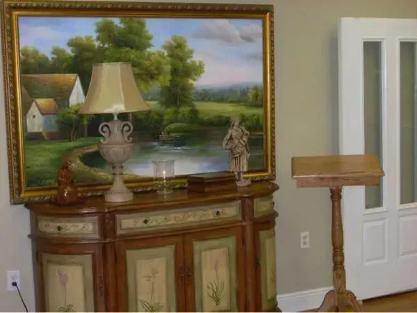 Living room with a large landscape painting, lamp-topped cabinet, small statue, and a wooden stand by a white door