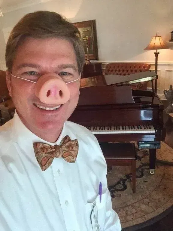 Man wearing a pig nose mask, bow tie, and glasses smiling in front of a piano in a warmly lit room