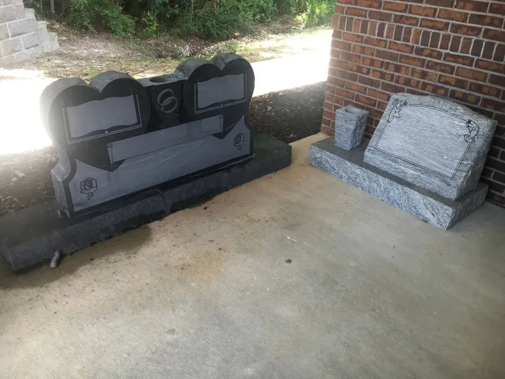 Two granite cemetery headstones beside a brick wall, one dark and one light gray.