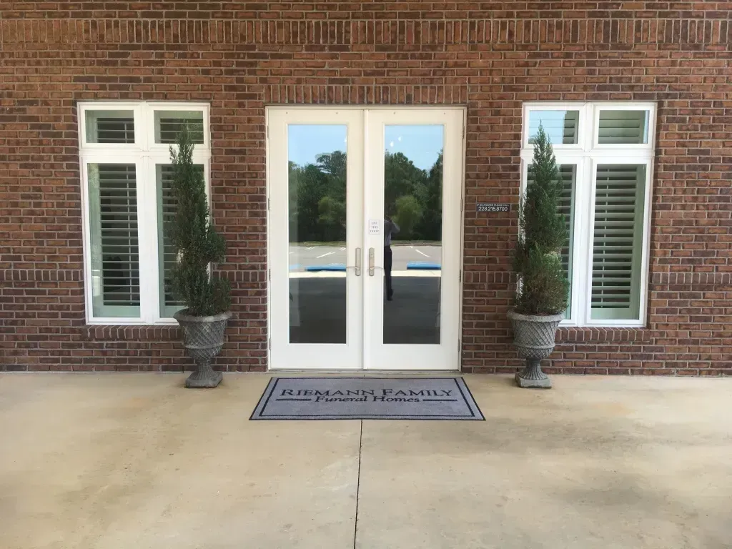 Brick building entrance with glass double doors, white-trimmed windows, and two potted topiary plants