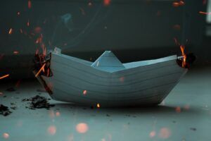 Paper boat afloat on calm water with glowing embers in a dark, moody scene