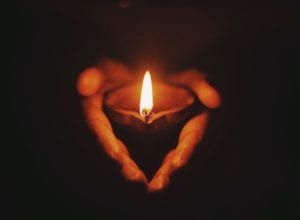 Hands cupped around a lit candle forming a heart shape in darkness