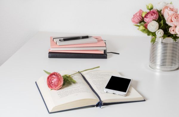 Open book with pink rose and phone on a white desk beside notebooks and flowers