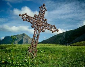 A rustic cross stands in a green field with mountains and blue sky in the background.