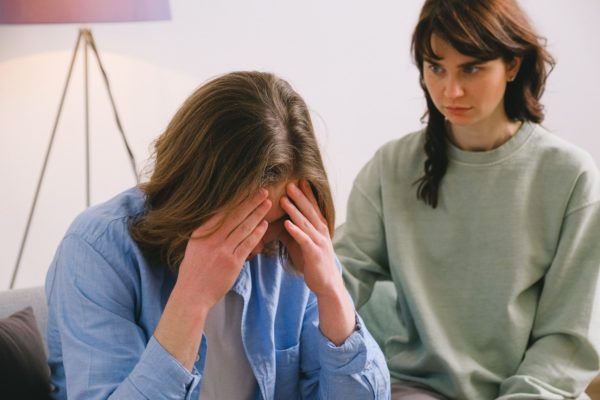 Two people sit indoors; one covers her face with her hands while the other looks on, concerned.