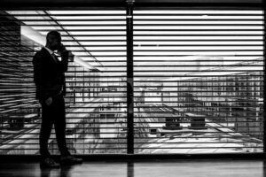 Silhouetted person by window blinds overlooking a cityscape in black and white
