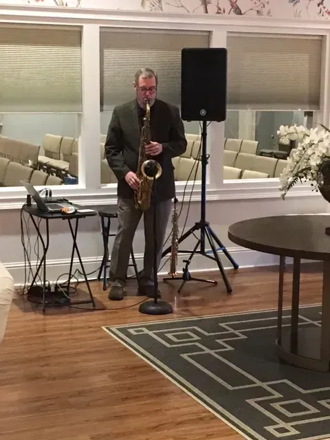 Musician playing saxophone indoors beside a speaker and microphone in a windowed room