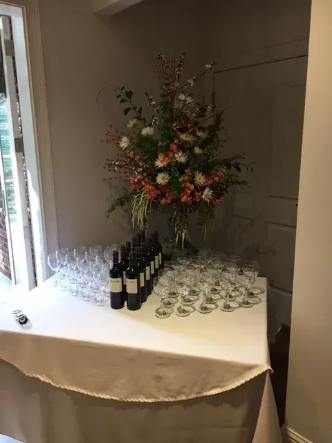 Wine bottles and glasses arranged on a table beside a tall floral centerpiece in a hallway.