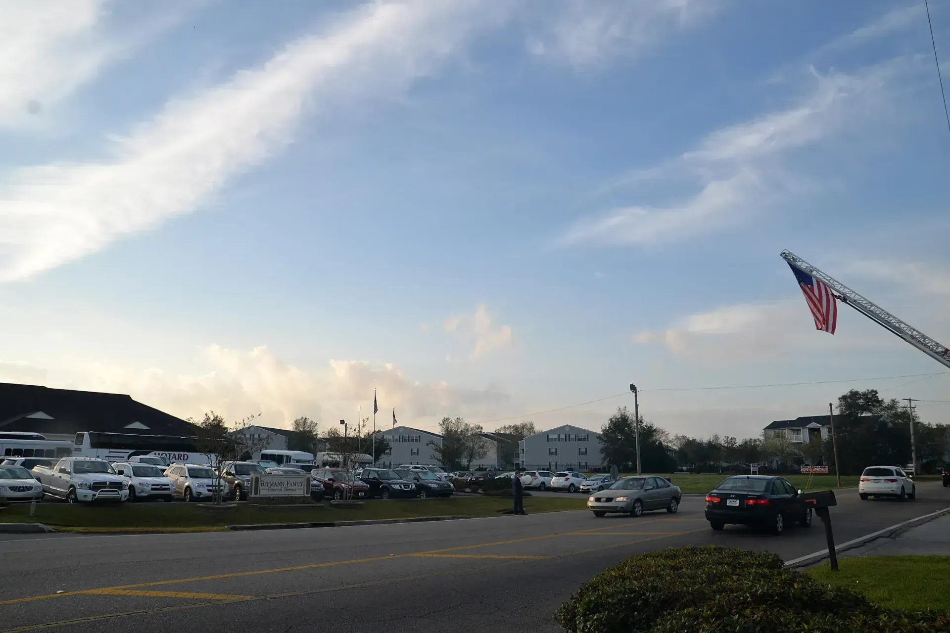 Parking lot at sunset with parked cars, trees, and a large American flag waving on the right.