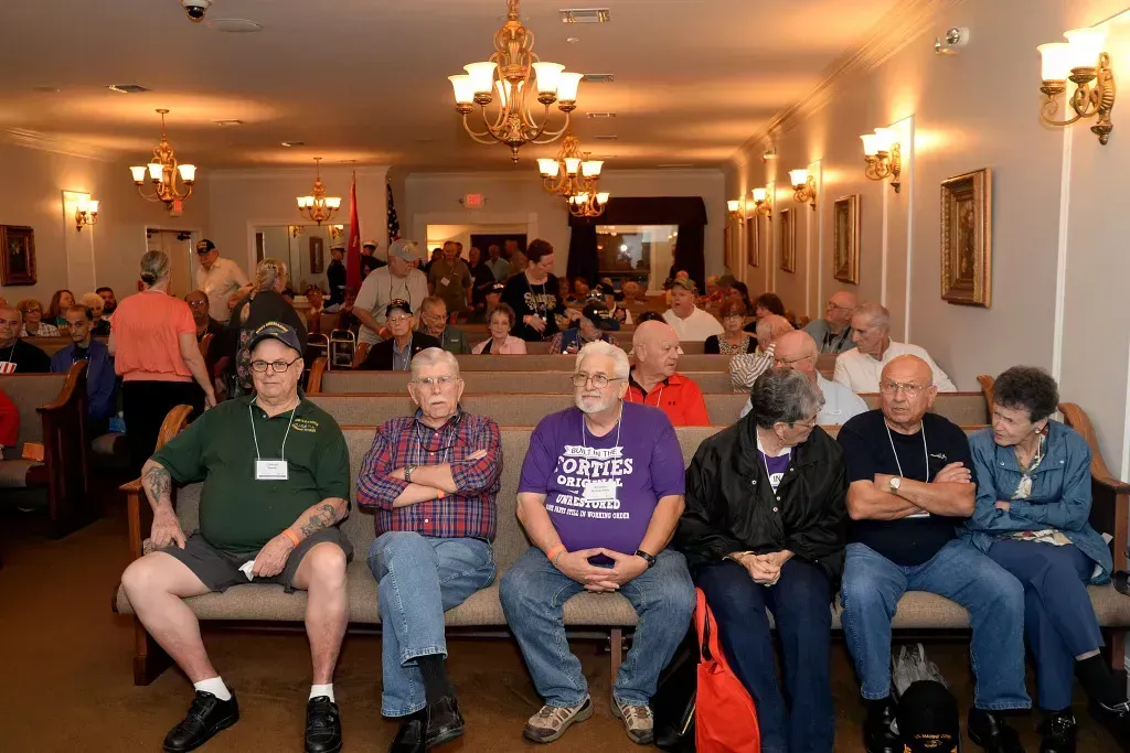 People seated in a warmly lit banquet hall, with a crowd gathered in the background.