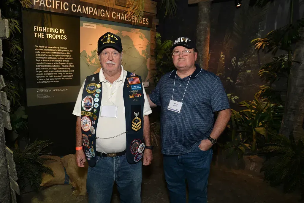 Two men wearing veteran hats pose in front of a Pacific campaign exhibit display.