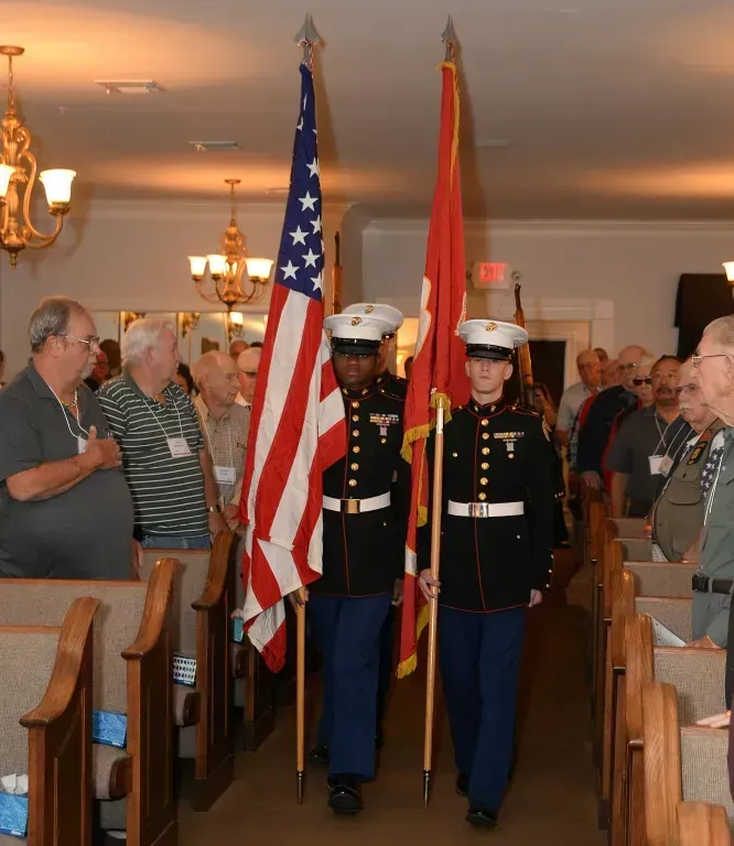 Color guard marching with U.S. and red flags through a crowded indoor hall