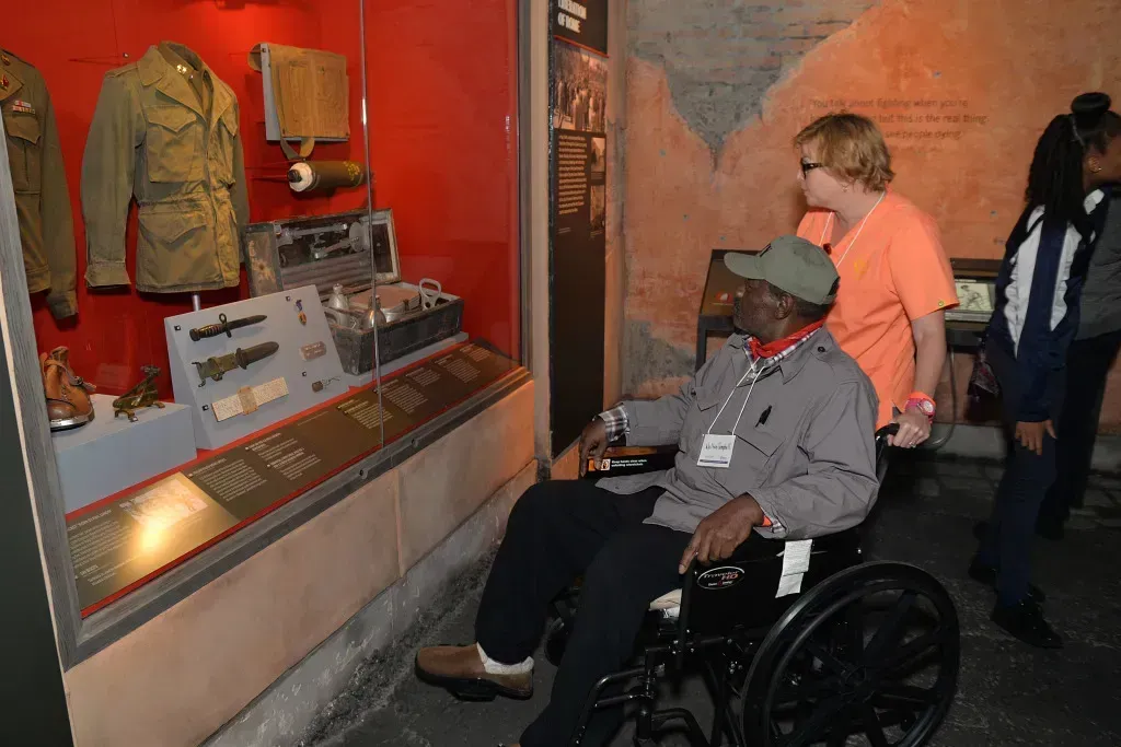Visitors in a museum viewing military uniforms and equipment behind glass, one seated in a wheelchair.