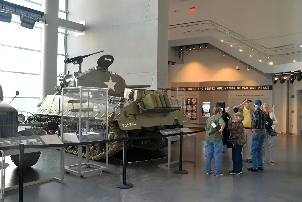 Museum exhibit with a green military tank and visitors standing nearby in a bright lobby