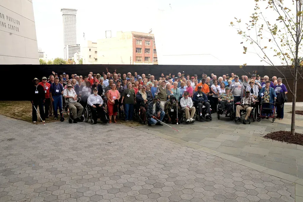 Large group posing outdoors in a courtyard, with a black wall and buildings in the background.