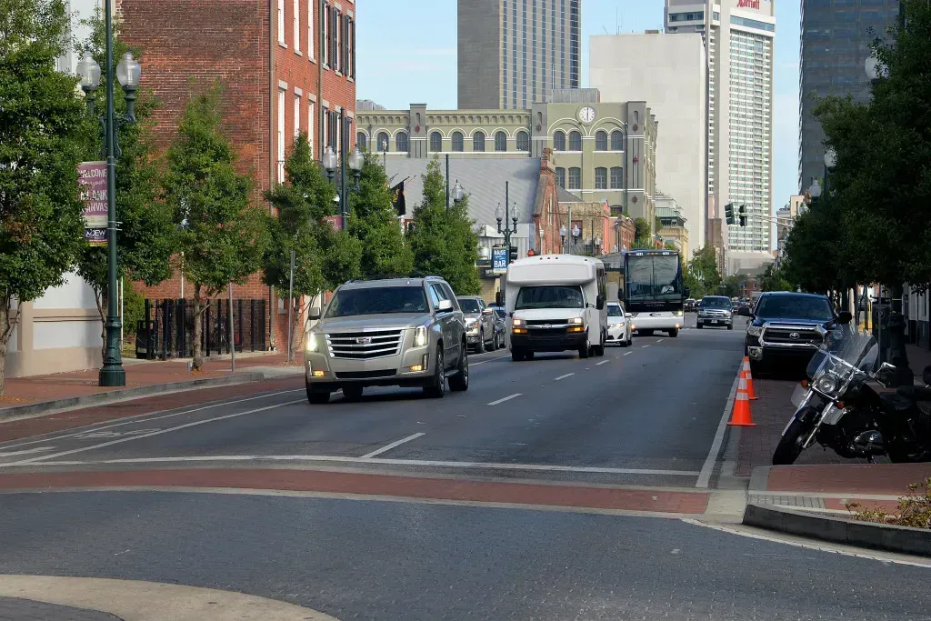 Downtown street with cars driving past brick and glass buildings, traffic cones along the curb.