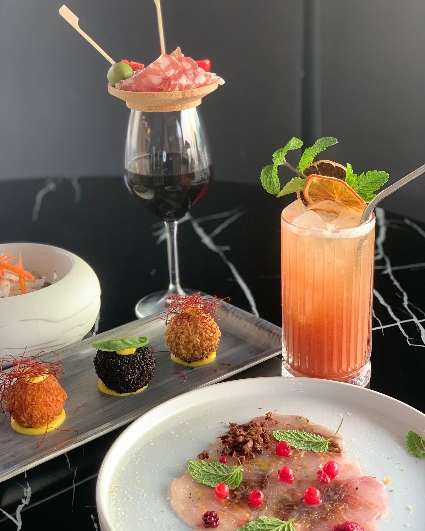Un elegante buffet di dessert e cocktail su un tavolo scuro, con dolci serviti su piatti, un calice di vino e una bevanda rosa alla menta.