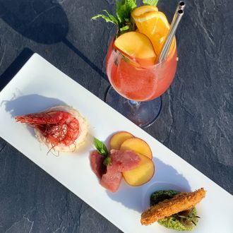 Piatto di antipasti gourmet con bocconcini di frutta e un cocktail rosa su un tavolo scuro, in piena luce del sole