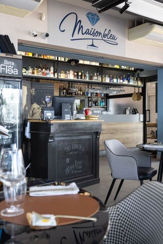 Accogliente bar-caffetteria con scaffali pieni di bottiglie, bancone con lavagna e sedie a scacchi sotto l'insegna