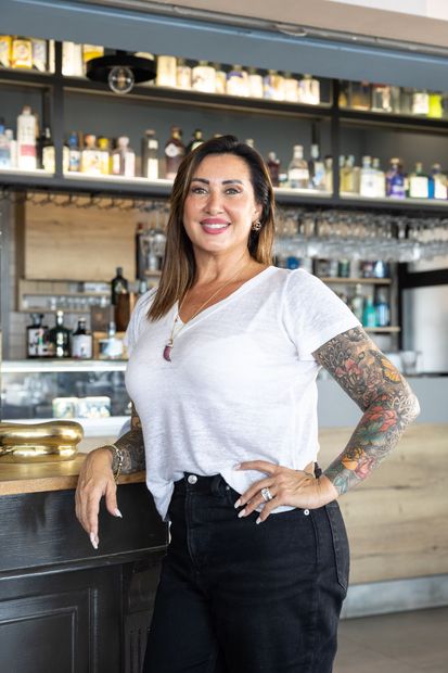 Una donna in piedi dietro un bancone, sorridente, con le braccia tatuate e scaffali di bottiglie sullo sfondo.