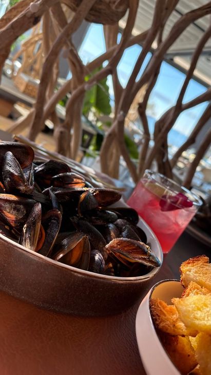 Una ciotola di cozze e patatine fritte su un tavolo in riva al mare, con una bevanda sullo sfondo.