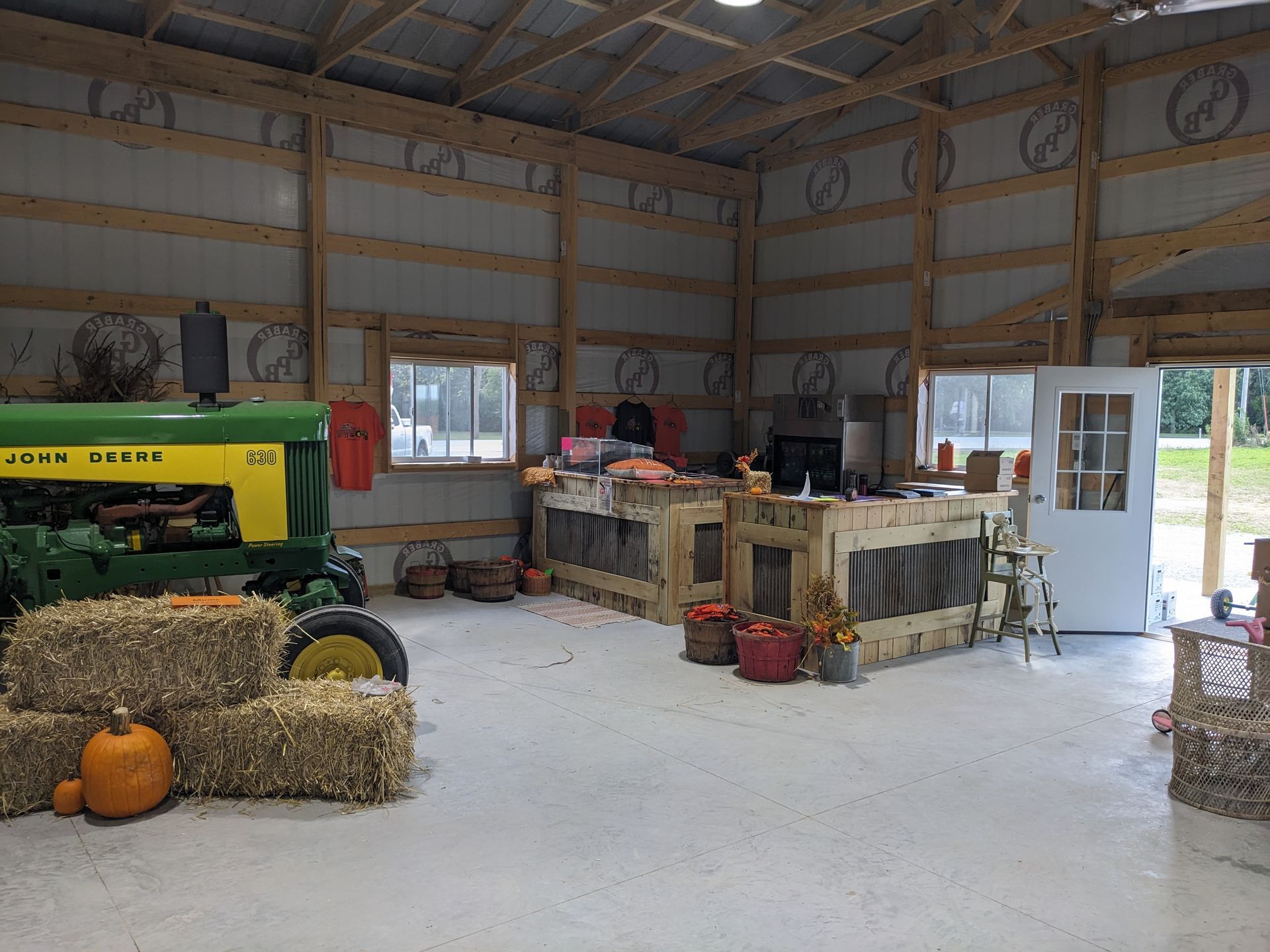 Farm barn interior with hay bales, pumpkins, and a green tractor near a window and exit.